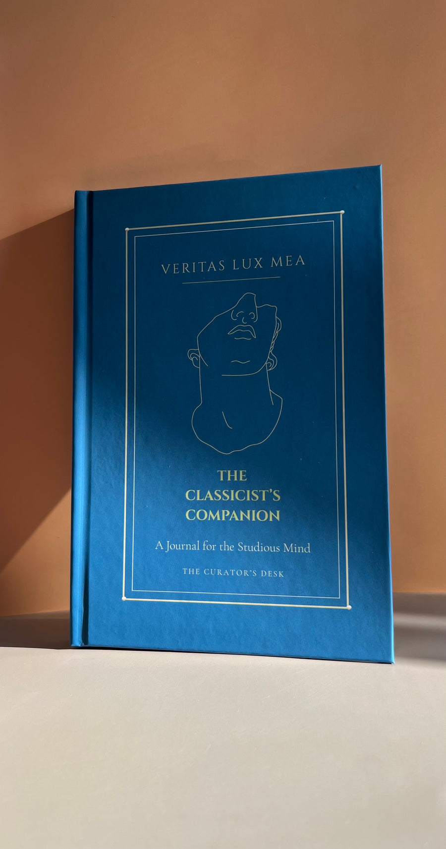 A hardcover journal titled 'The Classicist's Companion' by Veritatis Lux Mea, featuring a deep blue cover with gold foil lettering and a minimalist line drawing of a classical bust, set against a warm beige background.
